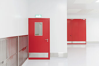 Schlüsselfertiger Hygieneraum/Reinraum von Plattenhardt+Wirth GmbH. Eine Edelstahlschleuse mit Wegbeschreibung dient in einem weiß gefliesten Flur als Zugangskontrolle. Alle Anschlüsse sind hygienisch und keimfrei ausgebildet. Die Türe ist rot neben einem Edelstahllüftungsgitter.